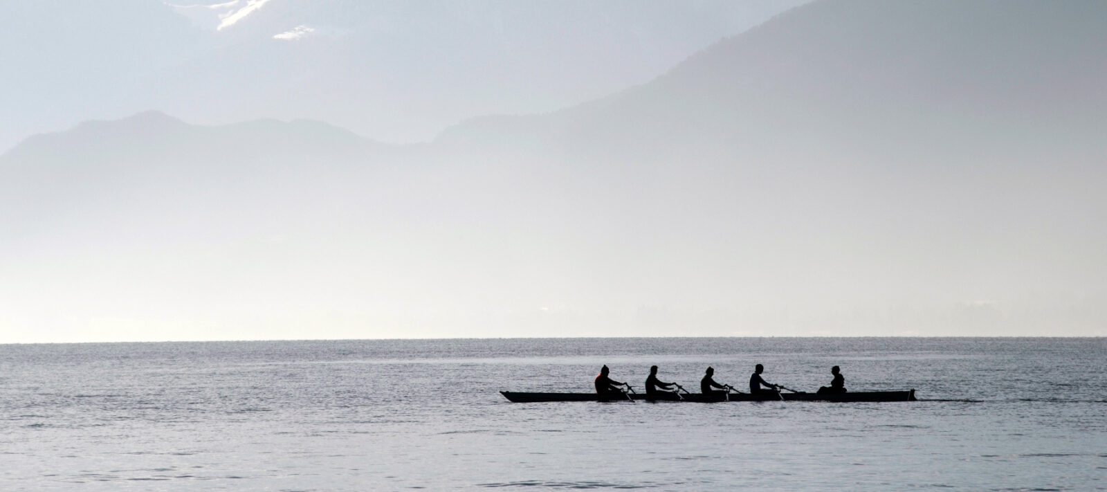 People rowing a long boat near mountains